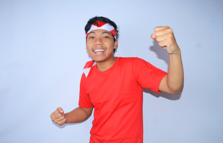portrait of excited indonesian young man raised his fist to celebrates his succeed or goal in indonesia independence day wearing red t shirt and headband. young man show yes gesture for his achievement and satisfactionの写真素材