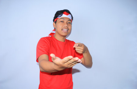 portrait of indonesian man smiling with left hand pointing to his open right hand palm wearing red t shirt and headband to celebrate indonesia independence day, looking to cameraの写真素材