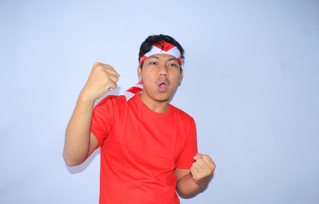 portrait of excited indonesian young man raised his fist to celebrates his succeed or goal in indonesia independence day wearing red t shirt and headband. young man show yes gesture for his achievement and satisfactionの写真素材