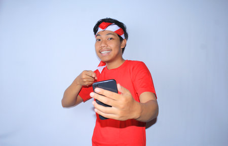 smiling indonesian young man pointing to his mobile phone with happy and excited face wearing red t shirt and headband isolated in whiteの写真素材