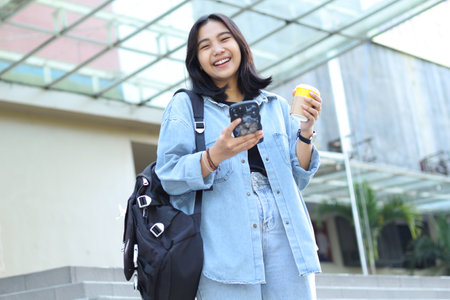 excited asian young woman is laughing while using app on smartphone and drinking coffee in outdoor stairs of a mallの写真素材