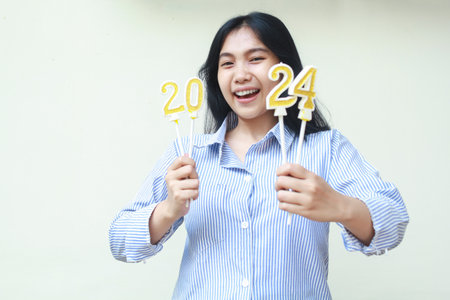 excited asian young woman coworker laughing with happiness celebrating new years eve by holding golden candles numbers 2024 wearing blue stripes shirt casual looking at camera isolated on white backgroundの写真素材