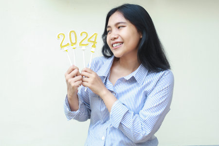 excited asian young business woman celebrating new years eve looking to golden candles numbers 2024 hold on hand wearing stripes blue shirt isolated on white backgroundの写真素材