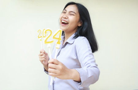 portrait of excited asian young woman coworker laughing at camera with closed eye and show 2024 figure candles lifting on hand wear grey formal shirt, isolated on white, new years eve conceptの写真素材