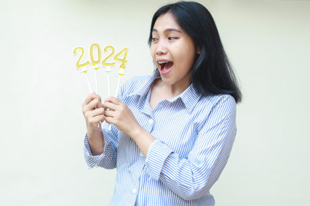surprised asian young woman screaming and looking aside to 2024 number candles hold on hand wearing casual blue stripes shirt, isolated on white, new years eve celebrationの写真素材