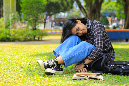 peaceful asian college student smiling and sitting in the lawn with book and backpackの写真素材
