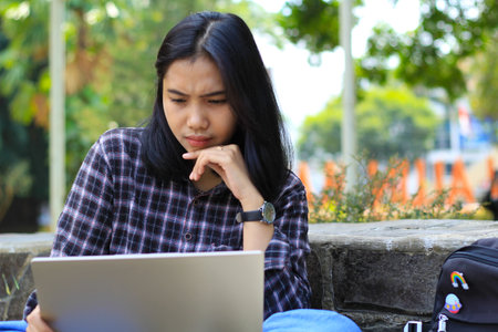 happy young asian woman focused using laptop  working remotely and browsing in social media in comfortable outdoors spaceの写真素材