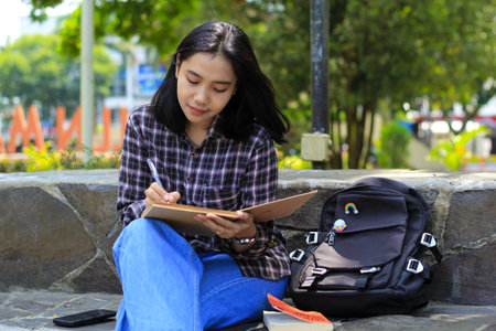 beautiful asian young woman college student focused writing on notebook and reading book in outdoors city parkの写真素材