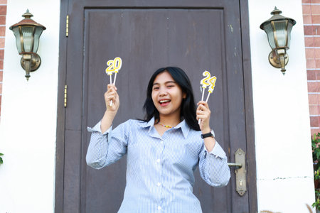 excited asian young woman raising hand holding 20 24 number candle and smiling to camera while standing in front of wooden door wearing blue stripes shirt casual, female coworker celebrate new yearの写真素材