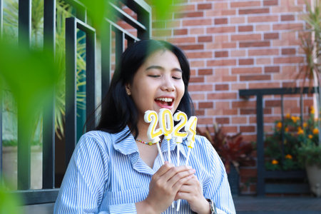 attractive young woman asian looking down to 2024 number candle holding on hand while standing over railing in outdoor with green leave framing, beautiful female standing over brick wall backgroundの写真素材