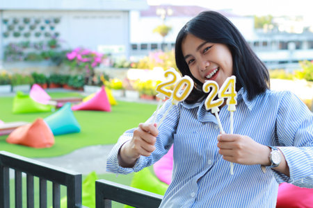 playful asian young woman looking at camera excited to show 2024 number candle holding on hand,...