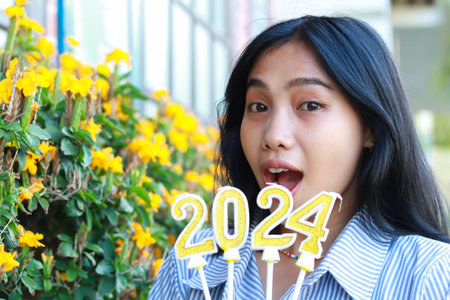 shocked asian young woman looking at camera with wow expression and screaming while holding 2024 number candle to celebrate new year eve in outdoor garden of vintage house with brick wallの写真素材