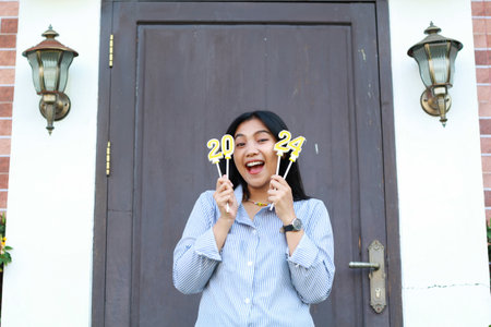 excited young woman celebrate new year eve with holding 2024 number candle, attractive female screaming to camera and standing over vintage house with wood doorの写真素材