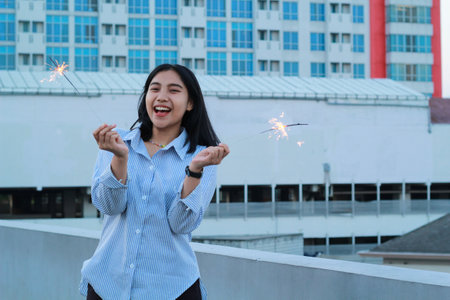 happy asian young business woman holding sparklers to celebrate new year eve standing in outdoor roof top with city building backgroundの写真素材