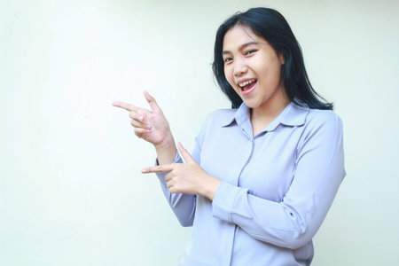 presenting asian young business woman excutive pointing finger to side looking to camera smiling with excitement, recommend product choices, wearing formal shirt isolated on white backgroundの写真素材