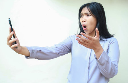 pretty asian young business woman shocked looking mobile phone hold on hands raising palm, questioning gesture, wearing formal shirt standing over isolated white backgroundの写真素材