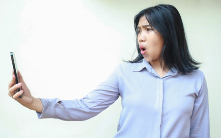 amazed asian young business woman using smartphone shocked with wow expression wearing formal shirt standing over isolated white backgroundの写真素材