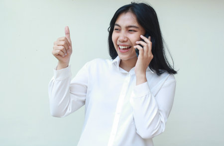 image of laughing asian young woman entrepreneur looking away giving thumbs up while speaking on smartphone wearing white shirt, good service gesture, standing isolatedの写真素材