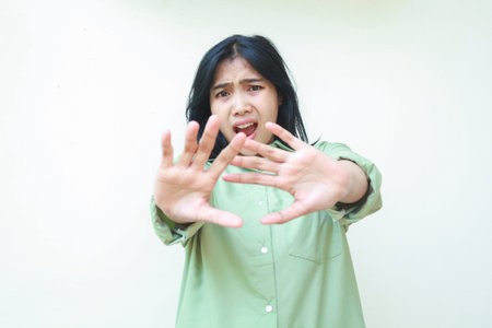 shocked dark hair asian woman reaching palms forward showing stop gesture wearing green oversized shirt looking at camera with scared face isolated on white backgroundの写真素材
