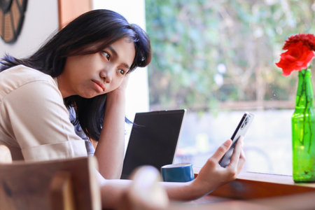 unhappy asian woman holding smartphone and grabbing her head feeling bored sitting near windows of coffe shop surfing social media, indoorの写真素材