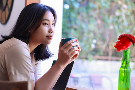 happy asian woman sitting in cafe drinkking coffee and working with laptop near window wearing fashionable clothes, smiling female remote worker holding mug in coffee shop look awayの写真素材