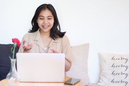 excited asian woman using laptop do online meeting with raising palms presenting gesture sitting on couch indoors wearing casual stylishの写真素材