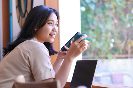 happy asian woman sit in cafe drink a cup of coffe and using smartphone with laptop on table wearing fashionable clothes, beautiful female work from coffee shop, relaxationの写真素材