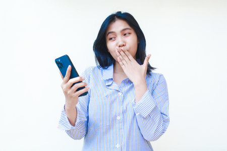 happy asian woman using mobile phone covering her mouth with surprised expression wearing striped shirt isolated on white backgroundの写真素材