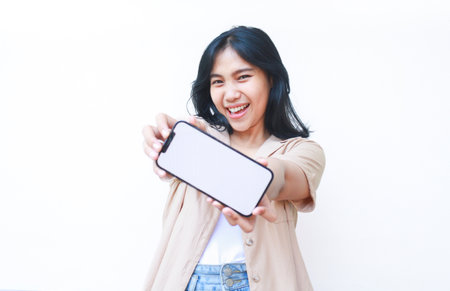 excited asian woman showing smartphone blank screen hold on hands to camera wearing casual brown clothes and jeans isolated on white backgroundの写真素材
