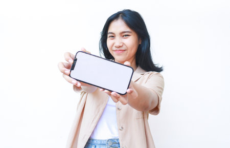 excited asian woman showing smartphone blank screen hold on hands to camera wearing casual brown clothes and jeans isolated on white backgroundの写真素材