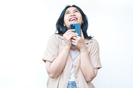 happy asian woman using smartphone laughing playful holding smartphone with raised hand wearing casual brown clothes isolated on white backgroundの写真素材