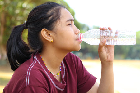 asian woman drinking a bottle of water while sitting under tree in outdoor feels tired and dehidratedの写真素材