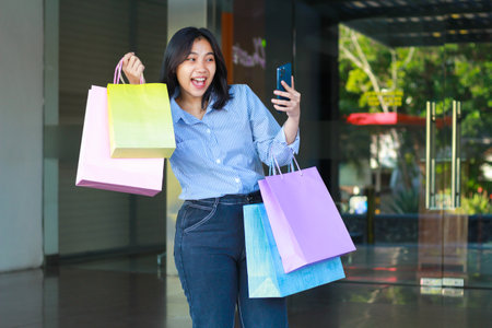 cheerful asian woman carrying shopping bag and holding smartphone to take selfie while walking in a mall outdoorsの写真素材