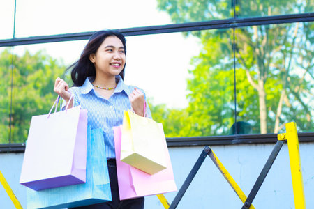happy beautiful asian woman holding shopping bag while walking in outdoors around mall building with excitementの写真素材