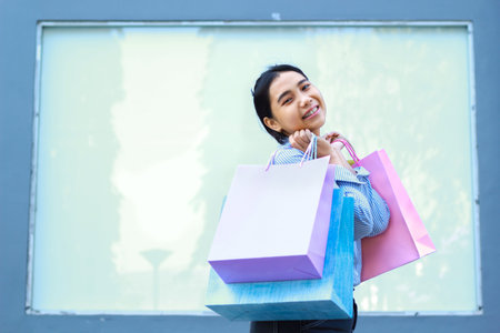 happy beautiful asian woman holding shopping bag with excitement standing in outdoor with empty billboard on backgroundの写真素材