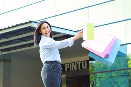 happy beautiful asian woman holding shopping bag while walking in outdoors around mall building with excitementの写真素材