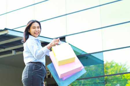 happy beautiful asian woman holding shopping bag while walking in outdoors around mall building with excitementの写真素材