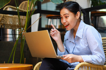 excited asian woman sitting in outdoor cafe with green plant interior using laptop working from anywhere, talking on computer have online meeting video chatの写真素材