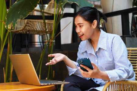 laughing asian woman digital nomad talking on laptop have video chat and online meeting with raising hands and holding smartphone working from outdoor cafeの写真素材