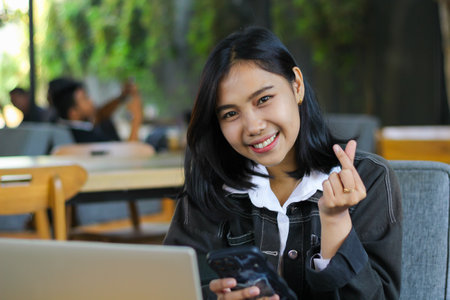 cheerful asian woman holding smartphone and show korean love signs while working with laptop...