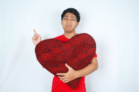 sad asian man hug heart shape pillow pointing finger above and looking at camera isolated on white backgroundの写真素材
