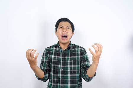 frustrated asian man wearing plaid flannel shirt raising hands and looking above with angry...