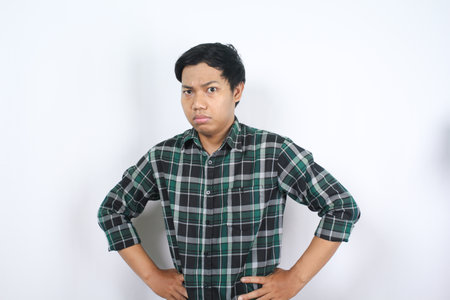 serious asian young adult man looking at camera with suspicion expression and hand on waist wearing casual shirt isolatedの写真素材