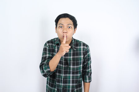 frightened asian man wearing casual plaid flannel shirt showing silent gesture with forefinger on...