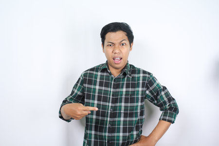 portrait of confused young asian man pointing finger to the middle with shocked and angry face wearing green plaid shirt isolated on white backgroundの写真素材