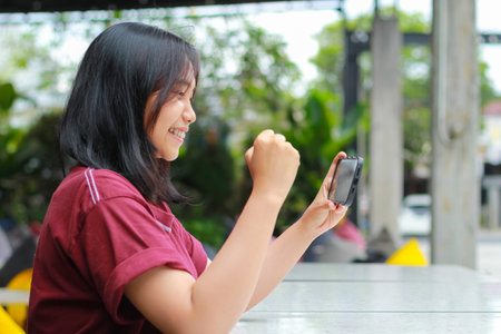 playful asian woman celebrating winning playing online game competition using smartphone with raising fist say yes gesture sitting in outdoor cafe, low angle viewの写真素材
