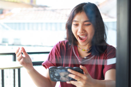 excited asian woman winning playing game online using smartphone in outdoor cafe shouting with open...
