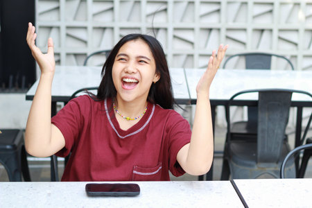 pretty asian woman sitting in cafe using smartphone and raising hand celebrating victory of online game tournamentの写真素材