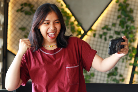 carefree asian woman celebrating winning playing online game tournament using smartphone with raised fist show yes gesture sitting in cafeの写真素材