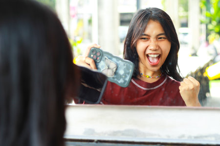 charming asian woman standing in front of mirror in cafe celebrating victory playing game online...
